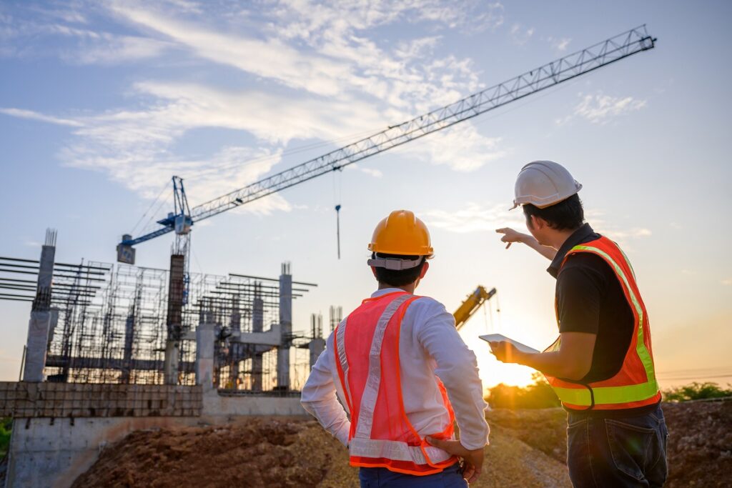 Construction site with two workers wearing safety vests and helmets, cranes and building framework in background at sunset